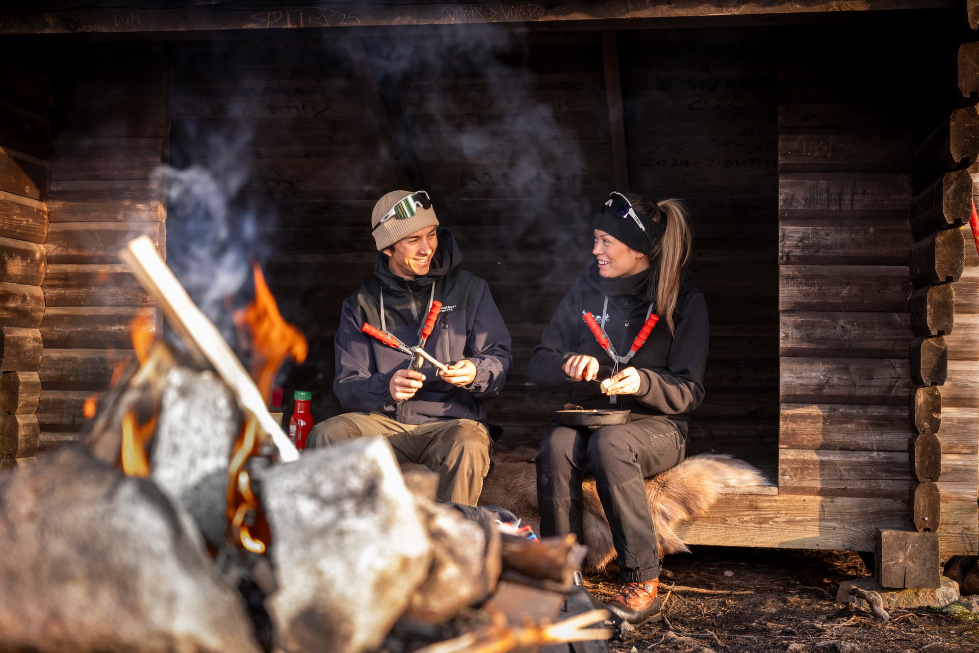Zwei Personen sitzen in einem Holzunterstand am Lagerfeuer, lächeln und bereiten Essen mit Spießen zu.