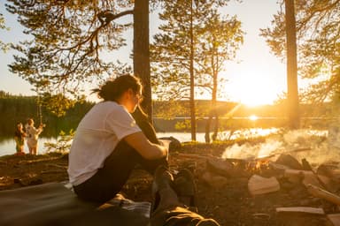 A group of people enjoying the sunshine while making food on a campfire during a canoe trip.