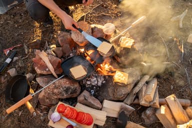 People making burgers over a campfire with all ingredients and tools scattered around the firepit.
