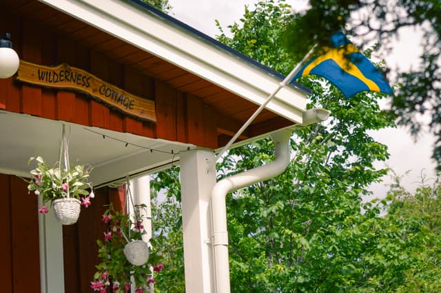 En träskylt med namnet 'Vildmarksstuga' och en svensk flagga som vajar i vinden.