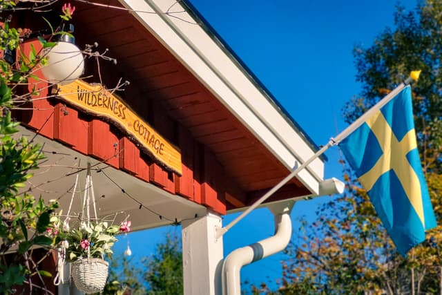 En träskylt med namnet 'Vildmarksstuga' i solljuset och en svensk flagga som vajar i vinden.