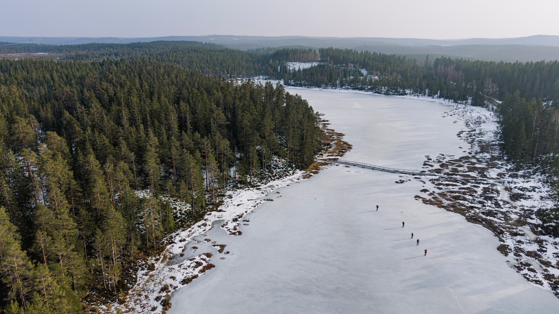 Luftaufnahme eines zugefrorenen Sees mit einer schmalen Holzbrücke und fünf Personen, die auf dem Eis neben einem dichten Kiefernwald laufen.