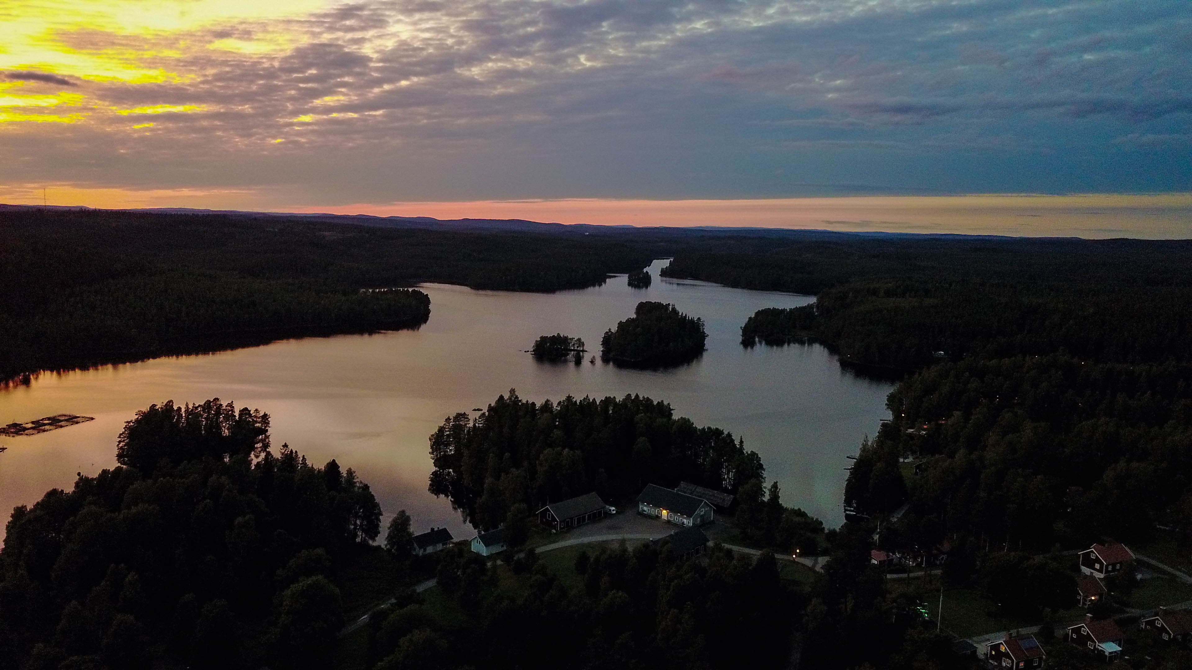 Una vista aerea di un lago tranquillo circondato da foreste e piccoli edifici al crepuscolo. Il cielo mostra una miscela di nuvole chiare e scure, con il tramonto che proietta un bagliore caldo sul paesaggio.