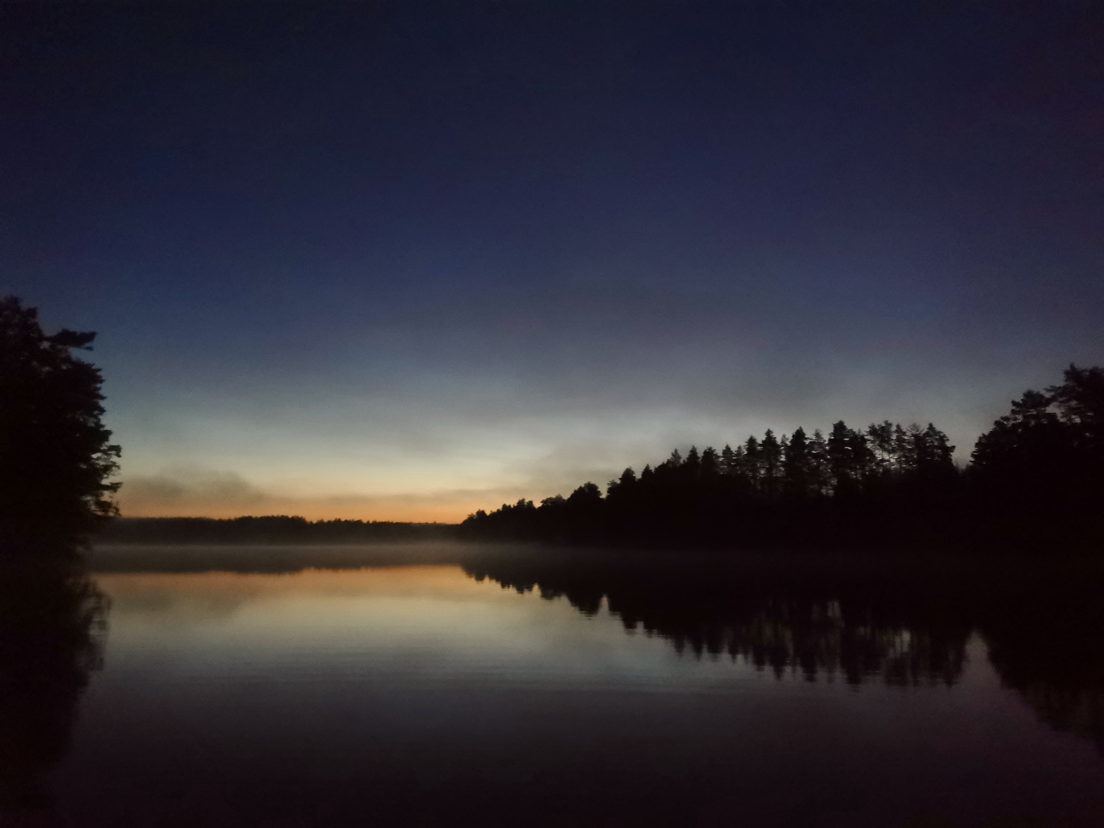 Un lago tranquillo al crepuscolo con il cielo che passa dalla luce al buio. La silhouette degli alberi si riflette nell'acqua calma, creando un'atmosfera pacifica e serena.