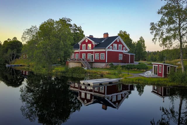 Från det stilla vattnet ser flodstugan vacker ut i solnedgången. Bastun, simområdet, däcket och den stora verandan speglas i vattnet.