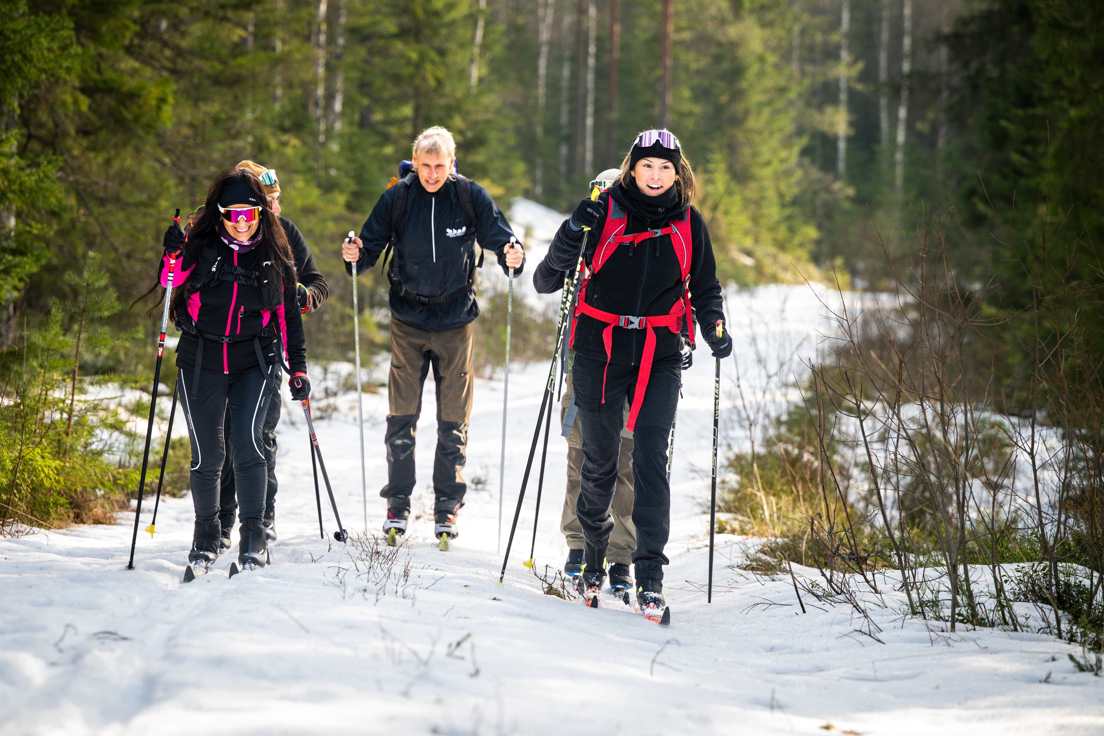 Fünf Personen skifahren auf einem verschneiten Waldpfad mit Stöcken, tragen Helme und Rucksäcke, Bäume säumen den Weg.