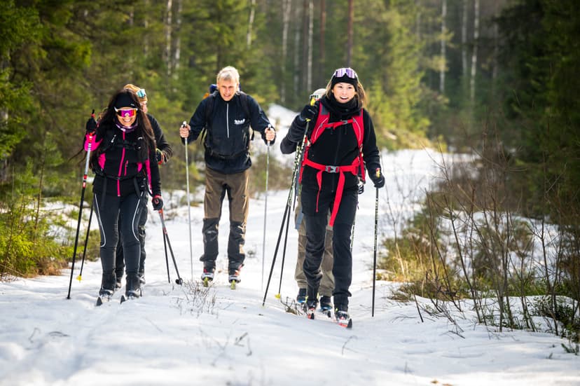 Cinq personnes skiant sur un sentier enneigé en forêt avec bâtons, portant casques et sacs à dos, arbres le long du chemin.