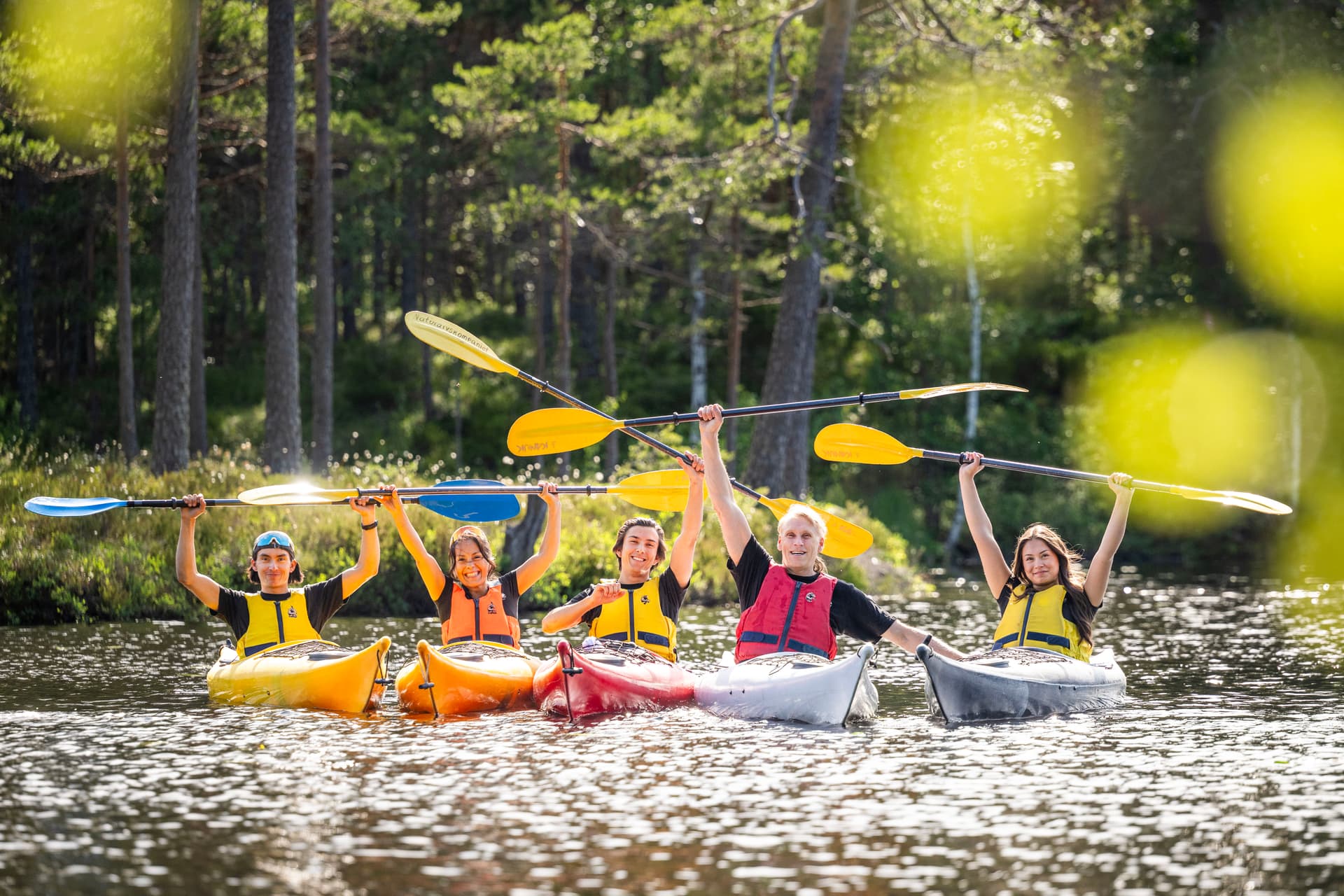 La famiglia Nilsson fa kayak in un lago con le pagaie sollevate in aria circondate da alberi verdi.