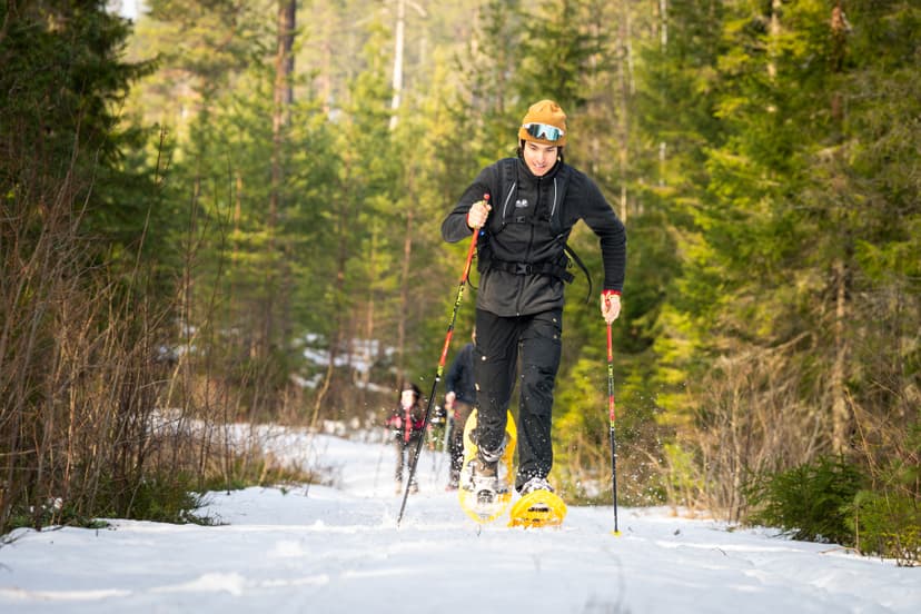 Une personne en raquettes avec bâtons sur un sentier enneigé en forêt, projetant de la neige, d'autres la suivent en arrière-plan.